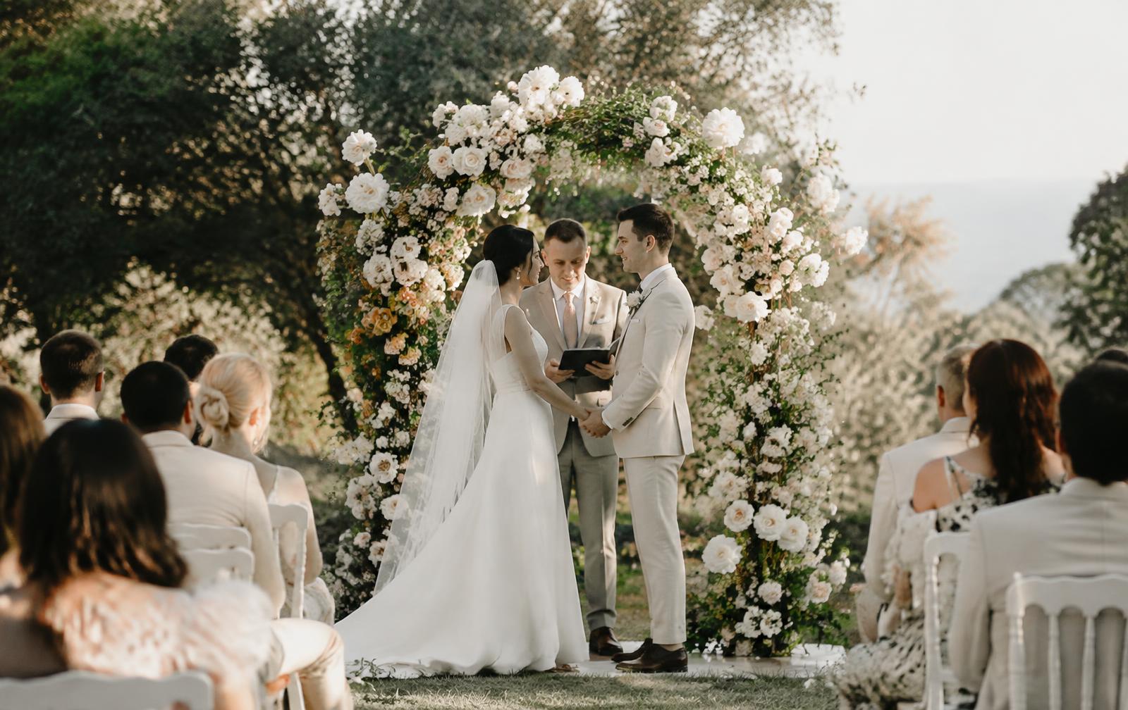 Couple exchanging vows beneath a white rose arch surrounded by guests